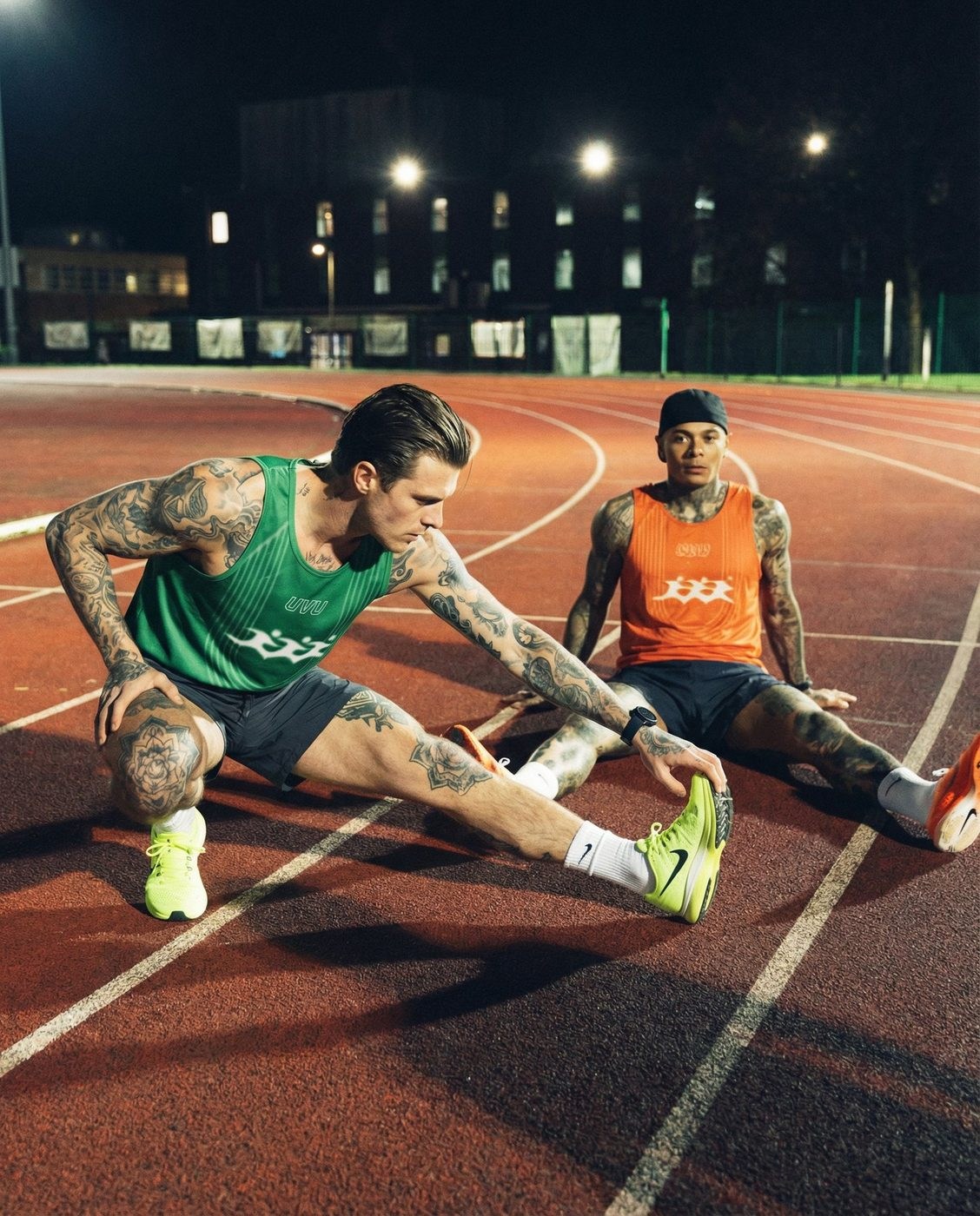 Dois atletas a alongar numa pista de atletismo à noite, com camisolas UVU